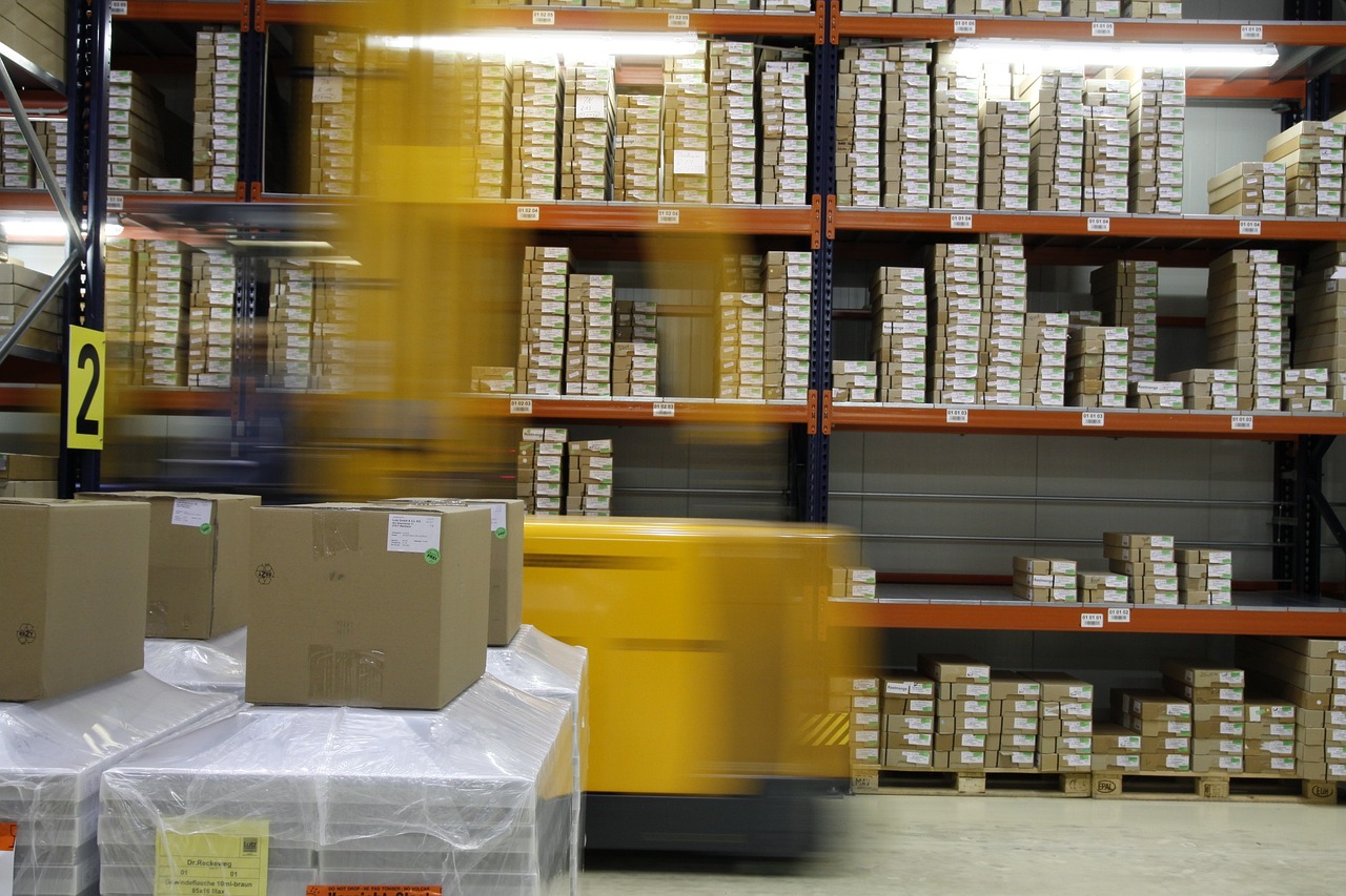 Warehouse shelving stocked with labelled cartons and a forklift passing in the foreground